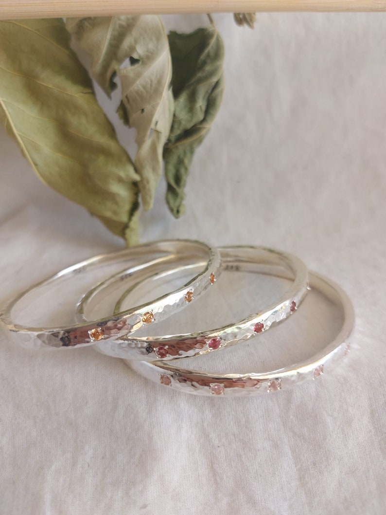 May include: Three silver bangles with hammered texture and small red and orange gemstones set into the metal.