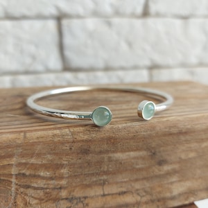 May include: A silver bangle bracelet with two light green gemstone accents. The bracelet is hammered and has a slightly open design.
