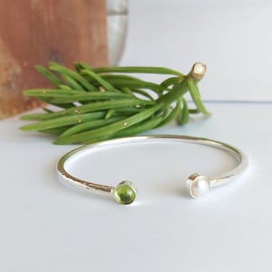 May include: A silver hammered bangle bracelet with a green peridot stone and a white pearl on either end.