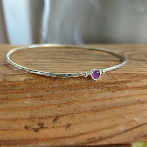 May include: A silver bangle bracelet with a small, round, purple amethyst gemstone set in the center.