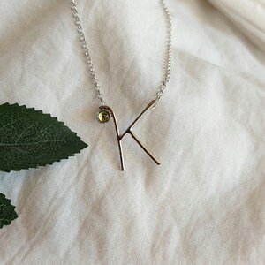 Op de afbeelding: Een zilveren ketting met een delicate letter K hanger en een kleine, ronde, gele edelsteen.