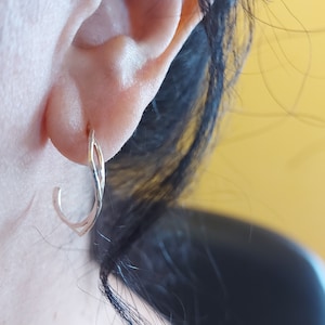 K&ouml;nnte beinhalten: Nahaufnahme eines silberfarbenen Ohrrings. Der Ohrring ist ein kleiner Reifen mit einem verdrehten Design. Der Ohrring wird am Ohr einer Person getragen, wobei Ohr und Haare im Hintergrund sichtbar sind. Der Hintergrund ist einfarbig gelb.
