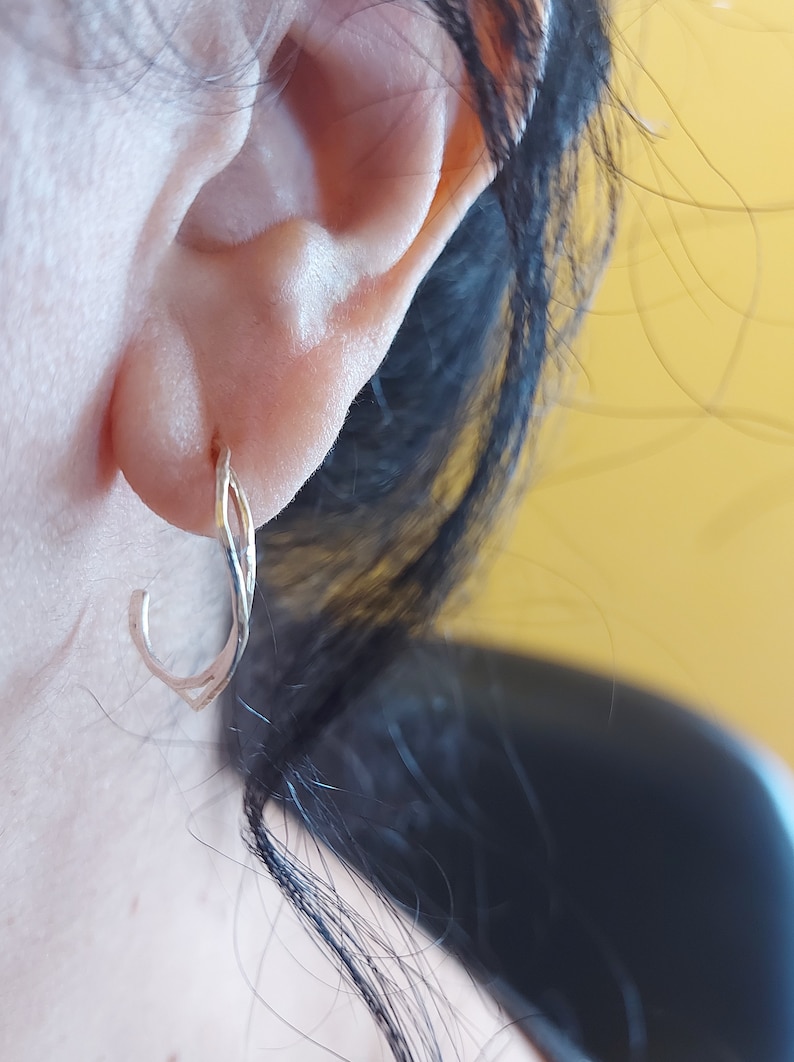 K&ouml;nnte beinhalten: Nahaufnahme eines silberfarbenen Ohrrings. Der Ohrring ist ein kleiner Reifen mit einem verdrehten Design. Der Ohrring wird am Ohr einer Person getragen, wobei Ohr und Haare im Hintergrund sichtbar sind. Der Hintergrund ist einfarbig gelb.