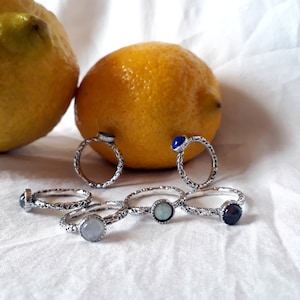 以下が含まれることがあります： 異なる色の宝石が付いた銀の指輪が6つ。指輪は互いに積み重ねられ、白い表面の上に置かれています。