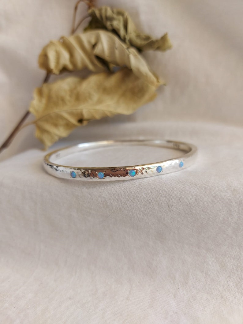 May include: A silver bangle bracelet with a hammered texture and four small blue opal stones set in a row.