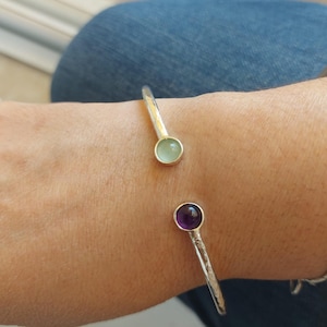 May include: A silver bangle bracelet with a hammered finish and two gemstone accents. The bracelet features a light green stone on one side and a purple stone on the other.