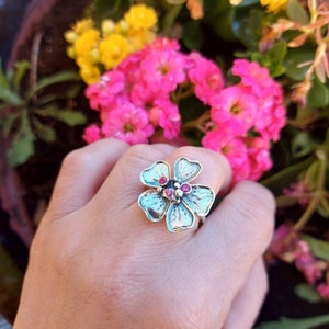 May include: A silver ring with a flower design featuring pink gemstones. The ring is on a hand against a background of pink and yellow flowers.