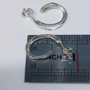 K&ouml;nnte beinhalten: Zwei silberne Creolen mit strukturierter Oberfl&auml;che. Die Ohrringe werden auf einem Lineal pr&auml;sentiert, die Reifen messen etwa 2,5 cm im Durchmesser. Die Ohrringe haben einen Stecker und einen Butterfly-Verschluss.