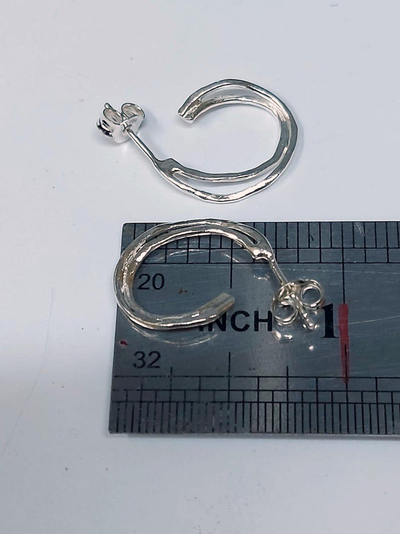 K&ouml;nnte beinhalten: Zwei silberne Creolen mit strukturierter Oberfl&auml;che. Die Ohrringe werden auf einem Lineal pr&auml;sentiert, die Reifen messen etwa 2,5 cm im Durchmesser. Die Ohrringe haben einen Stecker und einen Butterfly-Verschluss.