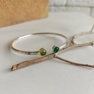 May include: A silver bangle bracelet with a hammered finish and two small round gemstones. One gemstone is green and the other is a light green color.