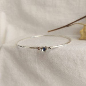 Puede incluir: Un brazalete de plata con un pequeño dije de flor con una piedra preciosa azul.