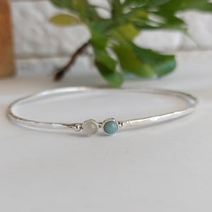 Pode incluir: Um bracelete de prata com duas pequenas pedras redondas, uma branca e outra azul claro.