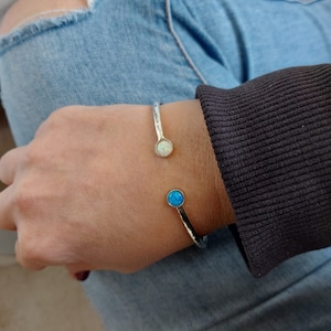 Pode incluir: Pulseira de punho prateada com textura martelada. A pulseira apresenta uma pedra de opala branca e uma pedra de opala azul. A pulseira é usada no pulso, com jeans azuis e uma camisa de manga comprida castanho escuro no fundo.