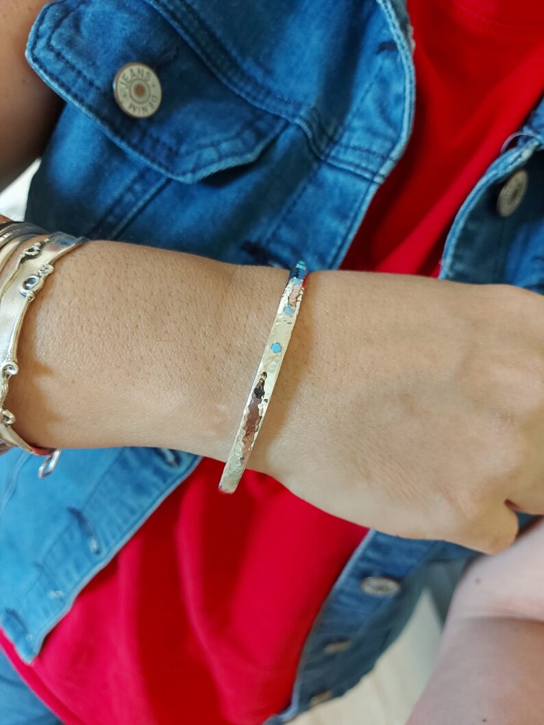 May include: A silver hammered bangle bracelet with small turquoise stones set into the metal. The bracelet is worn on a person's wrist.
