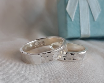 Puede incluir: Dos alianzas de boda plateadas martilladas, una ligeramente más grande que la otra, se muestran sobre una superficie de tela blanca. Las alianzas son lisas y tienen un acabado texturizado.
