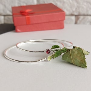 Op de afbeelding: Twee zilveren armbanden met gehammerde texturen. De armband bovenaan heeft een groene edelsteen, en de armband onderaan heeft een rode edelsteen.