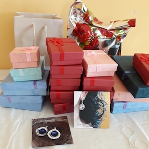 K&ouml;nnte beinhalten: Eine Sammlung von Geschenkboxen in verschiedenen Farben, darunter Rot, Rosa und Blau, mit passenden B&auml;ndern gebunden. Eine silberne Geschenkt&uuml;te und ein Geschenk mit floralem Design sind ebenfalls vorhanden. Schmuckkarten befinden sich im Vordergrund.
