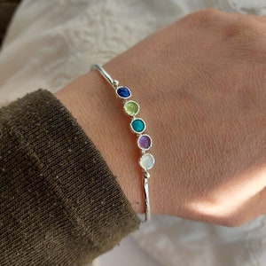 May include: A silver bangle bracelet with a row of colourful gemstones. The stones are blue, green, yellow, purple, and white.