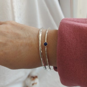 May include: Two silver hammered bangle bracelets with a small blue gemstone set in a gold bezel on one of the bracelets.