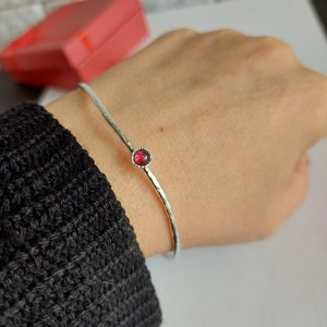 Puede incluir: Un brazalete de plata con acabado martillado y una sola piedra preciosa roja engastada en el centro.