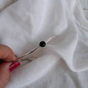 May include: A silver bangle bracelet with a black gemstone set in a silver bezel. The bracelet is on a white fabric background.