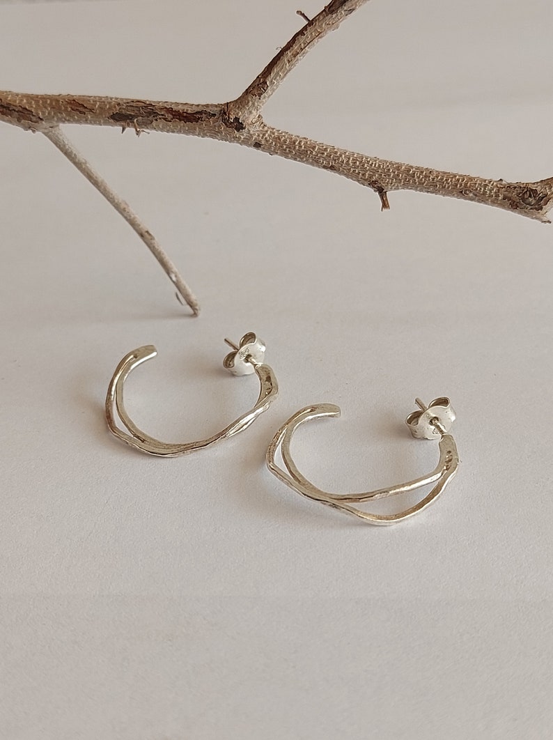 K&ouml;nnte beinhalten: Ein Paar silberne Creolen mit einem einzigartigen, strukturierten Design. Die Ohrringe haben eine doppellagige, offene Kreisform und werden mit Stecker- und Butterfly-Verschl&uuml;ssen befestigt. Ein Zweig liegt im Hintergrund.