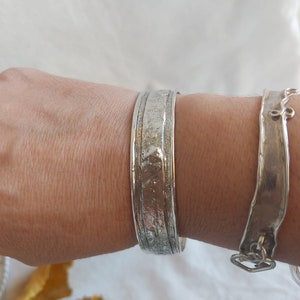 May include: A silver bracelet with a textured surface. The bracelet is wide and has a slightly rounded shape.