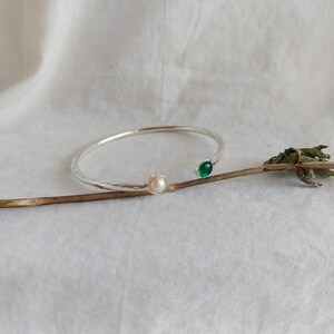 May include: A silver bangle bracelet with a white pearl and a green gemstone. The bracelet is on a white background.