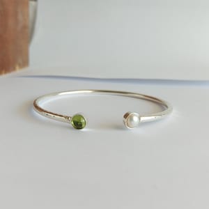 Può includere: Un braccialetto rigido in argento con finitura martellata. Un'estremità del braccialetto presenta una pietra preziosa verde, mentre l'altra estremità presenta una perla bianca.