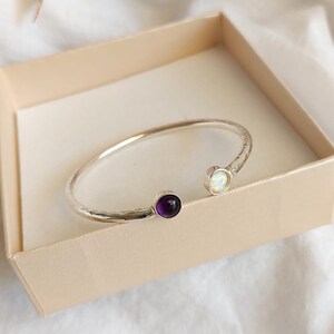 May include: A silver bangle bracelet with a hammered finish and two gemstones. One gemstone is a round, purple amethyst and the other is a round, white opal.