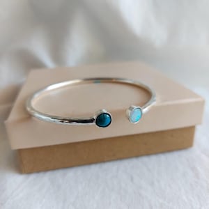 Può includere: Un bracciale a polsino argentato con una pietra turchese e un'opale. Il design aperto ne facilita l'indossatura. Il bracciale è esposto su una superficie dai toni neutri.