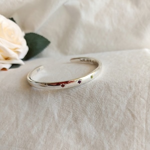 May include: Silver bangle bracelet with three small gemstones: purple, green, and red. The bracelet is engraved with the words "We are all unique".