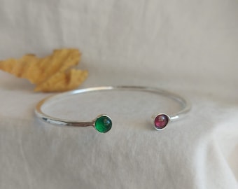 May include: A silver bangle bracelet with a green and a pink gemstone. The bracelet is open and has a hammered finish.