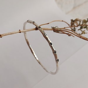 Może przedstawiać: Srebrna bransoletka typu obręcz z teksturowaną powierzchnią i serią małych, ciemnych kamieni szlachetnych. Bransoletka ma prosty, elegancki design i jest prezentowana na neutralnym tle z elementami suszonych roślin. Średnica: około 6.5 cm.