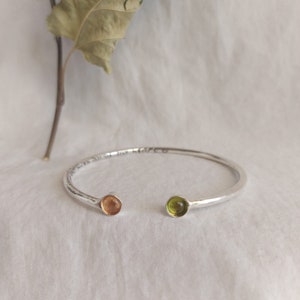 May include: Silver bangle bracelet with a hammered finish and two small round gemstones. One gemstone is orange and the other is green.