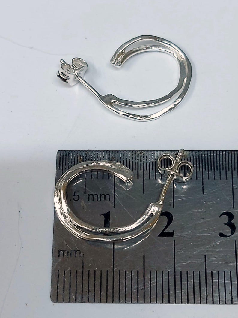 K&ouml;nnte beinhalten: Zwei silberfarbene Creolen. Jeder Ohrring hat einen Stecker und ein doppellagiges, C-f&ouml;rmiges Design. Die Ohrringe werden neben einem Lineal gezeigt, mit einem Durchmesser von etwa 1,9 cm.