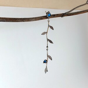 Puede incluir: Un pendiente plateado con un diseño de hojas y ramas. El pendiente presenta dos acentos de piedras preciosas azules y varias hojas pequeñas a lo largo del tallo. El pendiente cuelga de una rama marrón sobre un fondo blanco.