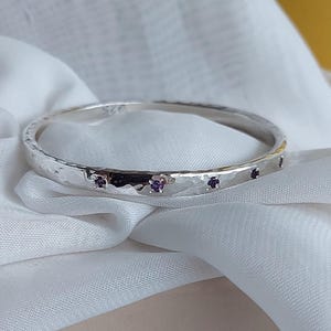 Może przedstawiać: Srebrna bransoletka z młotkowaną fakturą, inkrustowana małymi, okrągłymi, fioletowymi kamieniami szlachetnymi. Bransoletka jest prezentowana na białej, jedwabistej tkaninie, tworząc kontrast.