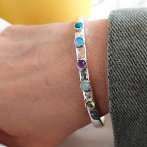May include: A silver-toned bangle bracelet with a hammered texture. The bracelet is adorned with six colorful gemstones: turquoise, blue, purple, aqua, and green. The bracelet is worn on a wrist.
