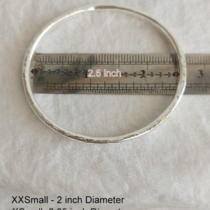 May include: A silver bangle bracelet with a hammered texture. The bracelet is measured against a ruler, showing a diameter of 2.5 inches. The image also includes text listing different sizes and their corresponding diameters: XXSmall - 2 inch Diameter, XSmall - 2.25 inch Diameter, Small - 2.5 inch Diameter, Medium - 2.75 inch Diameter, Large - 3 inch Diameter.