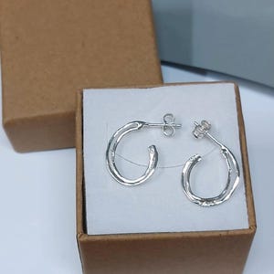 K&ouml;nnte beinhalten: Ein Paar silberne Creolen in einer kleinen, offenen, braunen Pappschachtel. Die Ohrringe sind halbmondf&ouml;rmig mit einem Steckerverschluss. Die Schachtel steht auf einer wei&szlig;en Oberfl&auml;che mit grauem Hintergrund.