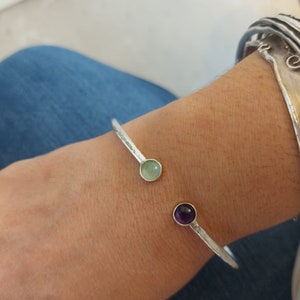 May include: A silver bangle bracelet with a light green and a purple gemstone. The bracelet is open and has a hammered texture.