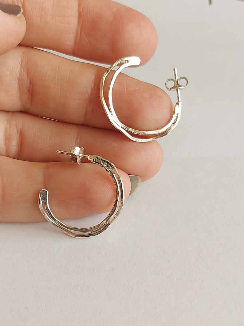 K&ouml;nnte beinhalten: Ein Paar silberne Creolen. Jede Ohrring hat ein zweischichtiges Design mit einer strukturierten Au&szlig;enschicht und einer glatten Innenschicht. Die Ohrringe werden vor einem neutralen Hintergrund pr&auml;sentiert.