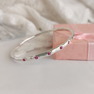 Puede incluir: Un brazalete de plata con textura martillada y piedras preciosas rosas engastadas en el borde.