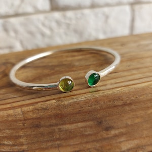 Puede incluir: Un brazalete de plata con acabado martillado y dos piedras redondas de vidrio, una verde y otra amarilla.
