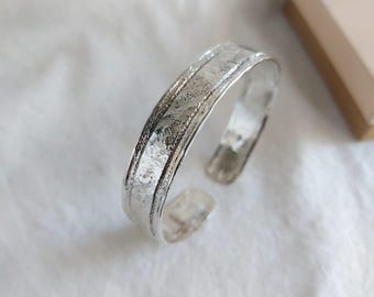 May include: A silver cuff bracelet with a textured surface. The open-ended bracelet has a wide band with a slightly hammered appearance. The jewelry is displayed on a white surface, with a small, tan box in the background.