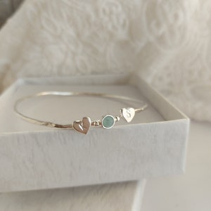 Può includere: Un braccialetto rigido in argento con una piccola pietra preziosa blu chiaro, due cuori d'argento e la lettera "S" incisa su uno dei cuori.