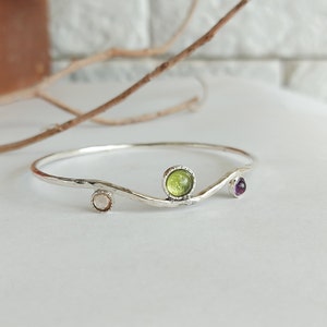 May include: A silver bangle bracelet with a wavy design and three gemstones. The gemstones are a light green, a light brown, and a purple.