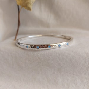 May include: A silver bangle bracelet with a hammered texture and five small blue opal stones set in the metal.