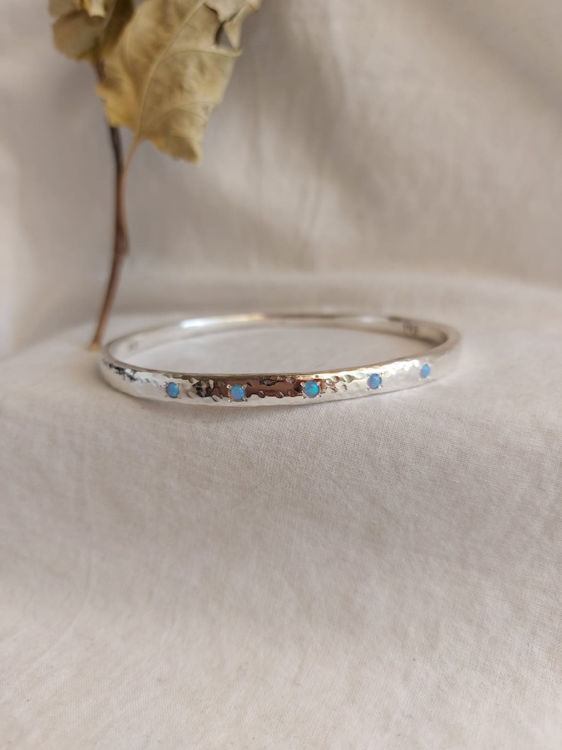 May include: A silver bangle bracelet with a hammered texture and five small blue opal stones set in the metal.